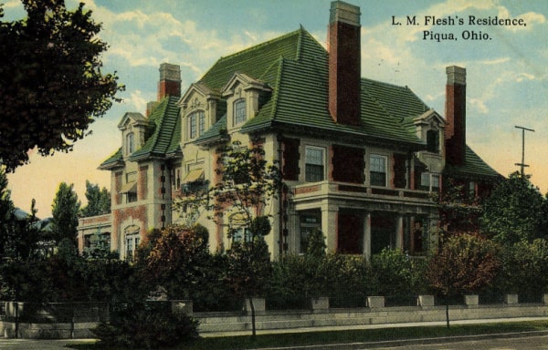 Historical postcard of L.M. Flesh's Residence, Piqua, Ohio. The postcard shows the mansion's northeast corner. There are shrubs around the home and a blue sky with some clouds behind it.