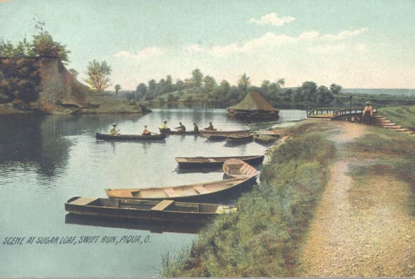 Historical postcard of Swift Run Lake. Several rowboats are on the lake, and two have people in them. There is a bridge on the right side of the postcard. Further in the background are trees and a blue sky with a few clouds. Text in the bottom left corner reads "Scene At Sugar Loaf, Swift Run, Piqua, O."