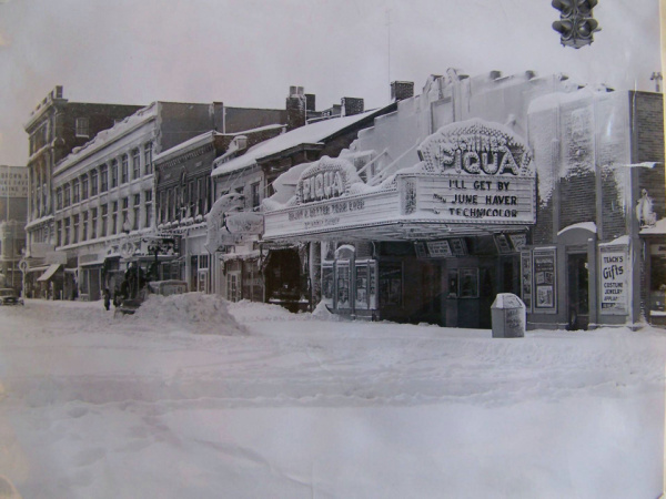 Image Description: A historical black and white photograph of Mainstreet in Piqua in the 1950s. The street is covered with snow, a plow can be seen and there are a few people o the sidewalk. The Schine's Piqua Theater is in the forefront of the photo. 