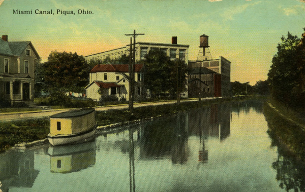 Historical postcard of the Miami Erie Canal. Along the left bank of the canal is a street, houses and industrial buildings. A small boat sits on the canal and the right bank is lined with trees.