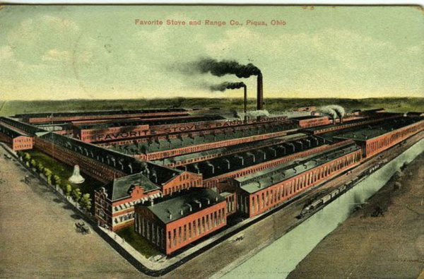 Historical postcard of the Favorite Stove and Range Co., in Piqua Ohio. The factory buildings are a deep brick red color, there is a water fountain in front of the building and black smoke coming from smokestacks.