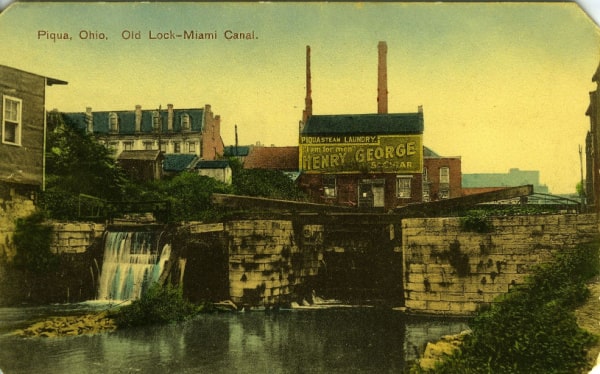 Historical postcard of the Piqua, Ohio Old Lock-Miami Canal. The image features the lock releasing water. The sky is pale blue/yellow and brick buildings are visible. Trees and shrubs grow along the canal and near the buildings. An advertisement for Piqua Steam Laundry is painted on a brick building.