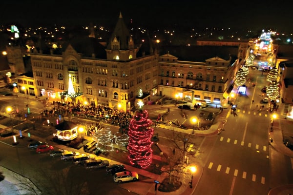 A modern, full-color postcard of downtown Piqua during the holiday season. The postcard is an aerial photograph of downtown at night. There are lights on the trees and the gazebo on the public square. People are gathered in front of the Fort Piqua Plaza, which appears to glow.