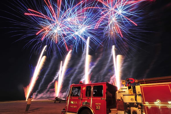 A modern, full-color postcard of red, blue, and white fireworks on a dark sky. In the background a small plane is visible. In the forefront is a red Piqua Fire Department truck.
